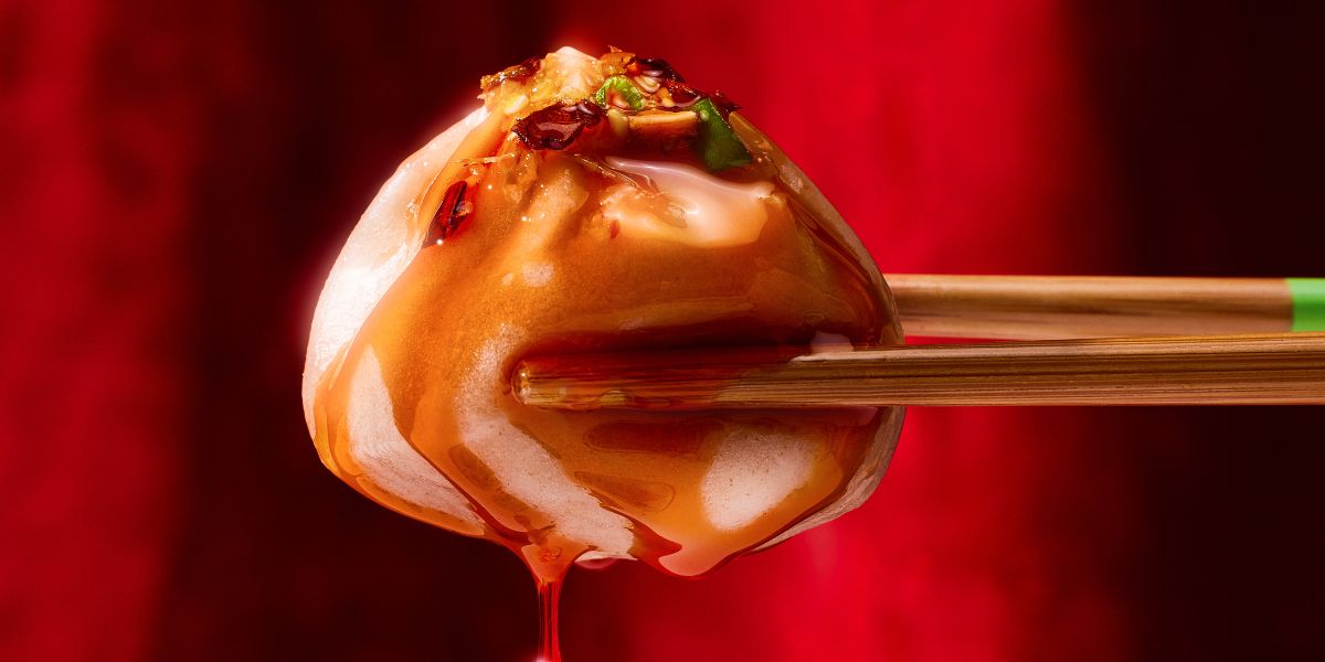 Close-up of a soup dumpling held in chopsticks, showing chili oil and dripping broth against a red background