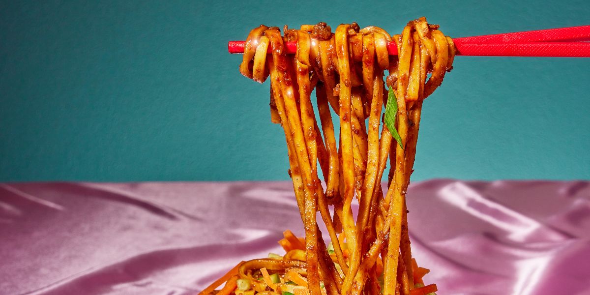 Close-up of spicy Chinese noodles lifted by red chopsticks showing chili oil coating and scallions against a teal background and pink satin surface