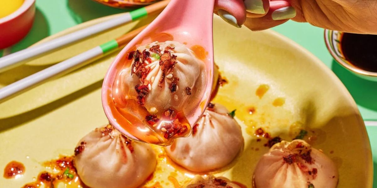 Soup dumpling on a pink spoon dripping chili oil above a yellow plate of dumplings with chopsticks nearby