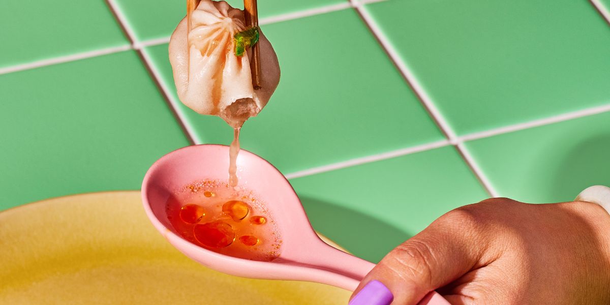 Soup dumpling held with chopsticks dripping broth into a pink spoon with chili oil against a green tiled background