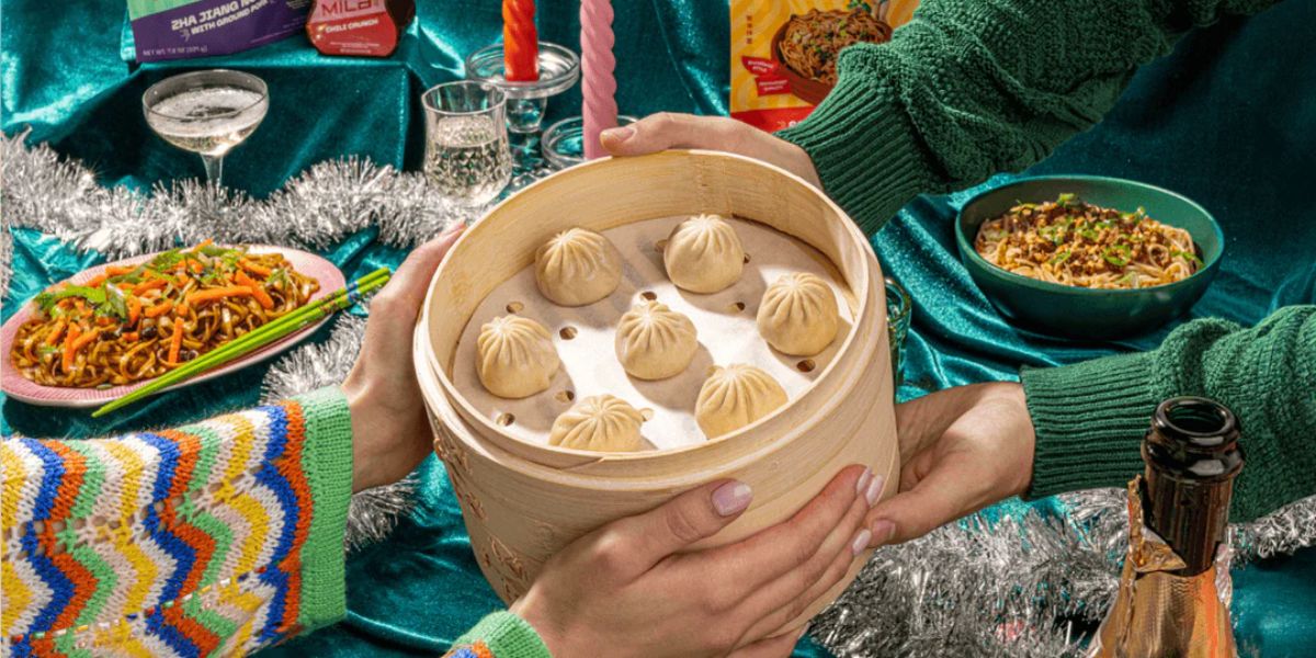 Two people passing a bamboo steamer of eight soup dumplings across a festive table with bowls of noodles, dipping sauces, and drinks