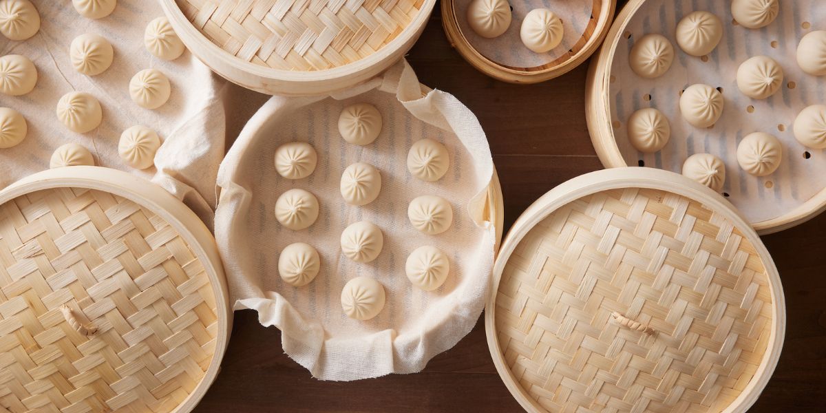 Rows of uncooked xiao long bao (soup dumplings) in bamboo steamers lined with cloth on a wooden table