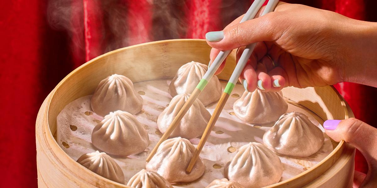 Hand using chopsticks to lift a steaming soup dumpling from a bamboo steamer with red cloth background