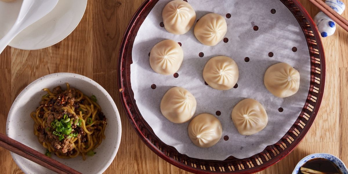 Top-down view of steamed soup dumplings in a bamboo steamer with a bowl of noodles, chopsticks, and dipping sauce