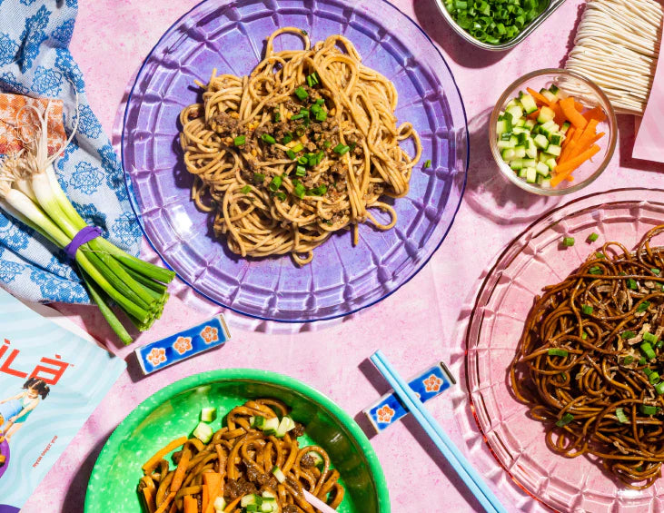 Overhead of three bowls of Chinese noodles with scallions, chopsticks, and a small bowl of chopped vegetables
