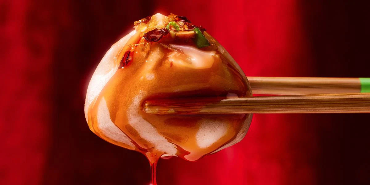 Close-up of xiao long bao soup dumpling held in chopsticks, glossy sauce dripping and chili flakes against a red background