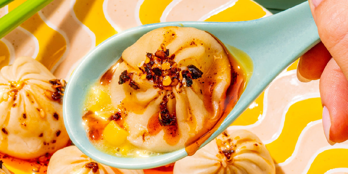 Close-up of a soup dumpling on a pale blue spoon showcasing steaming broth and chili oil with other dumplings on a yellow patterned plate