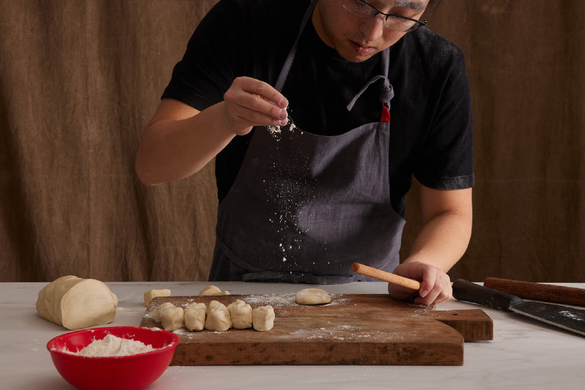 Home cook sprinkling flour over dough pieces while preparing dumpling wrappers on a wooden board with a rolling pin and red bowl of flour
