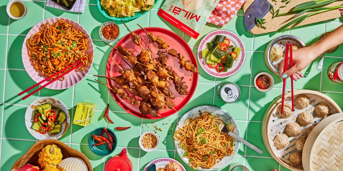 Chinese comfort food spread: soup dumplings, skewers, noodles and pickles on a green tiled table, hand picking a dumpling