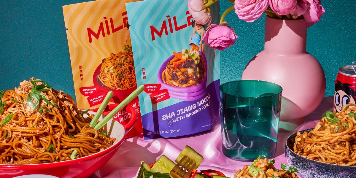 MìLà noodle packets and two bowls of prepared Zha jiang noodles on a pink tabletop with a green glass and pink vase