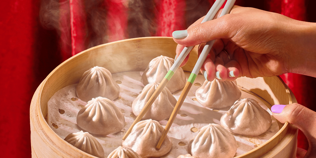 Close-up of soup dumplings in bamboo steamer showing pleated tops and rising steam, hands with chopsticks and red curtain