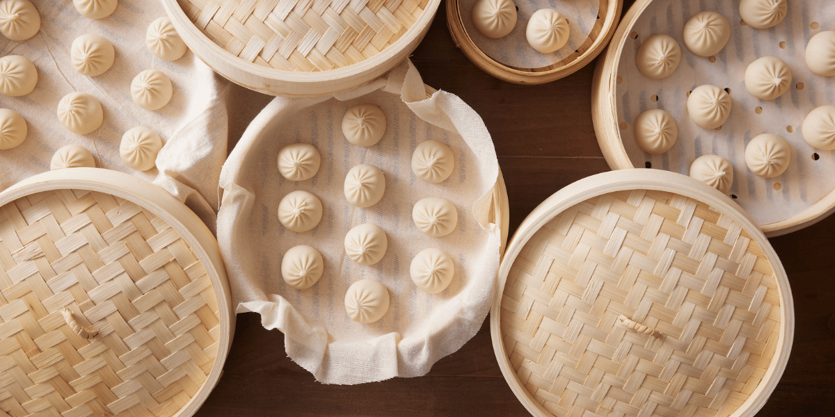 Bamboo steamer baskets holding rows of soup dumplings on cloth liners, arranged on a wooden table ready for steaming