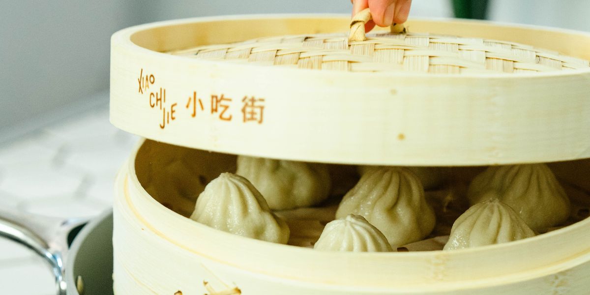 Xiao long bao steaming in a bamboo basket as a hand lifts the lid, close-up of soup dumplings