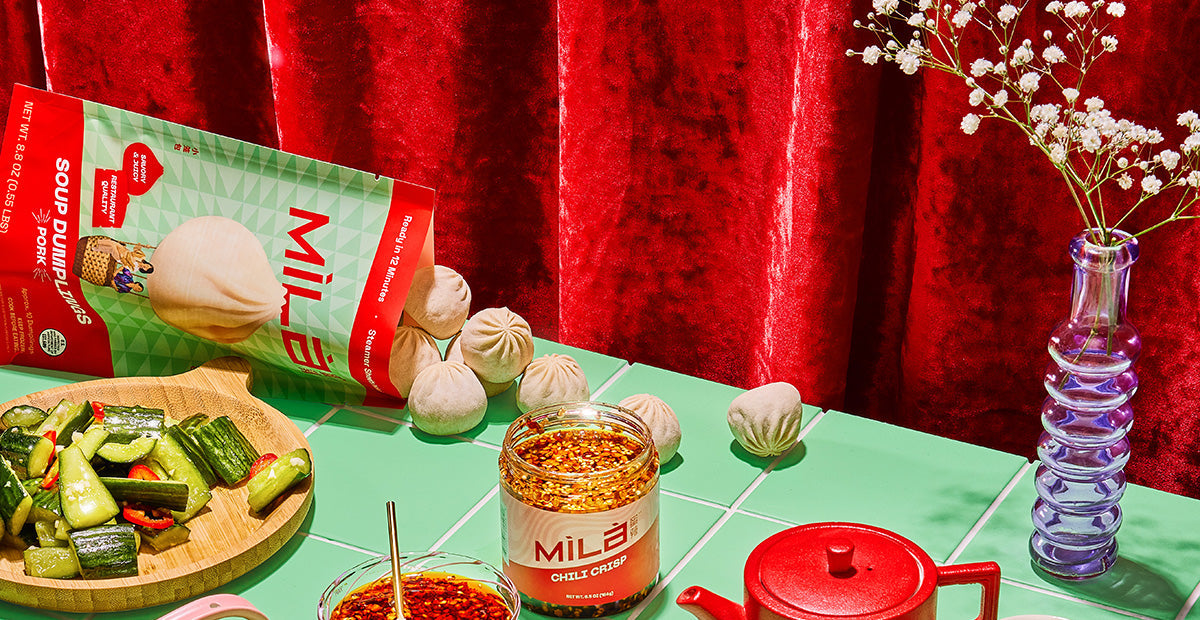 MìLà dumpling package and chili crisp jar on a green tiled table with cucumber salad and a red teapot against a red velvet curtain