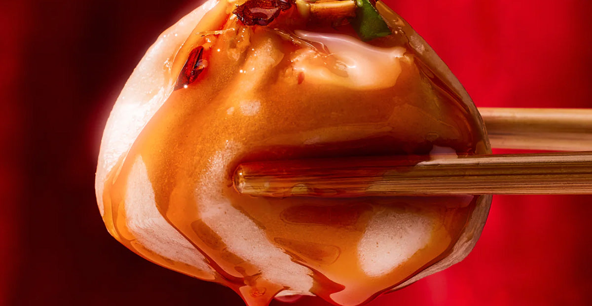 Chopsticks holding a steamed dumpling draped in glossy chili oil and crunchy garlic flakes against a red background