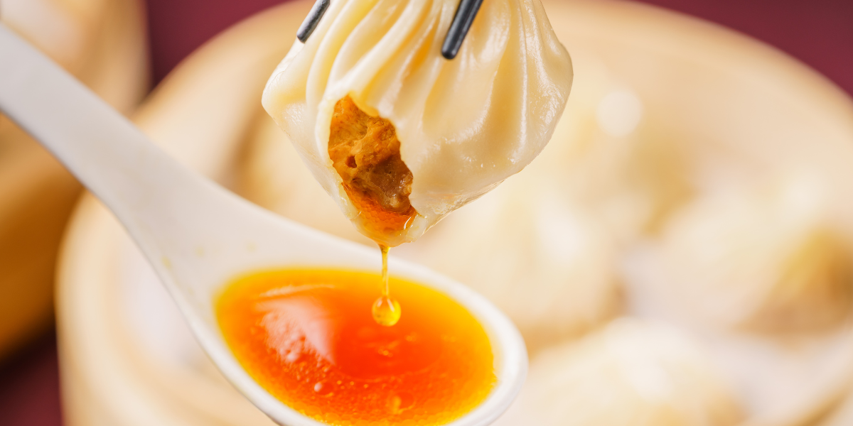 Soup dumpling lifted by chopsticks as hot broth drips into a white spoon over a blurred bamboo steamer background