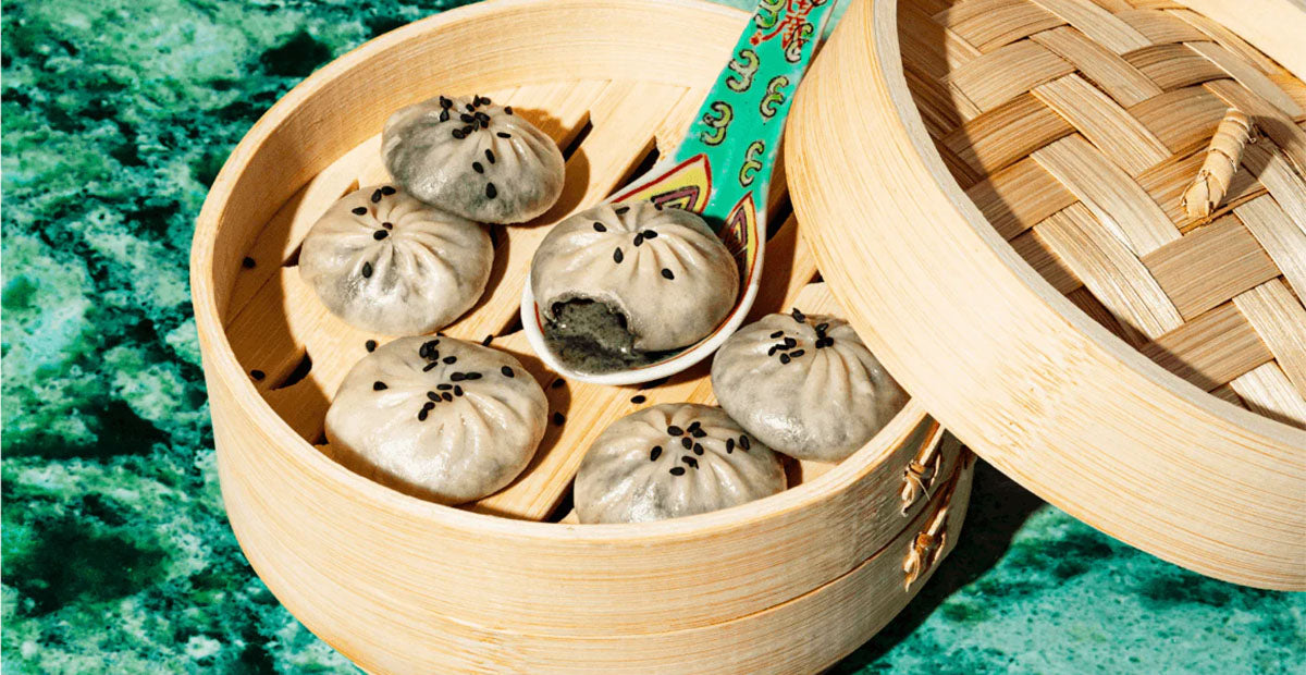 Top view of black sesame chocolate dumplings in steamer, one opened showing black filling and sesame seeds on green marble