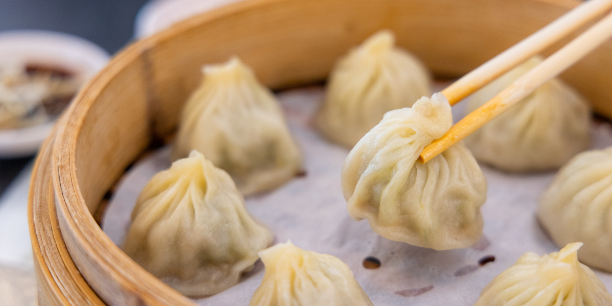 Close-up of steamed soup dumplings in a bamboo steamer, chopsticks lifting one to show pleated wrapper and filling