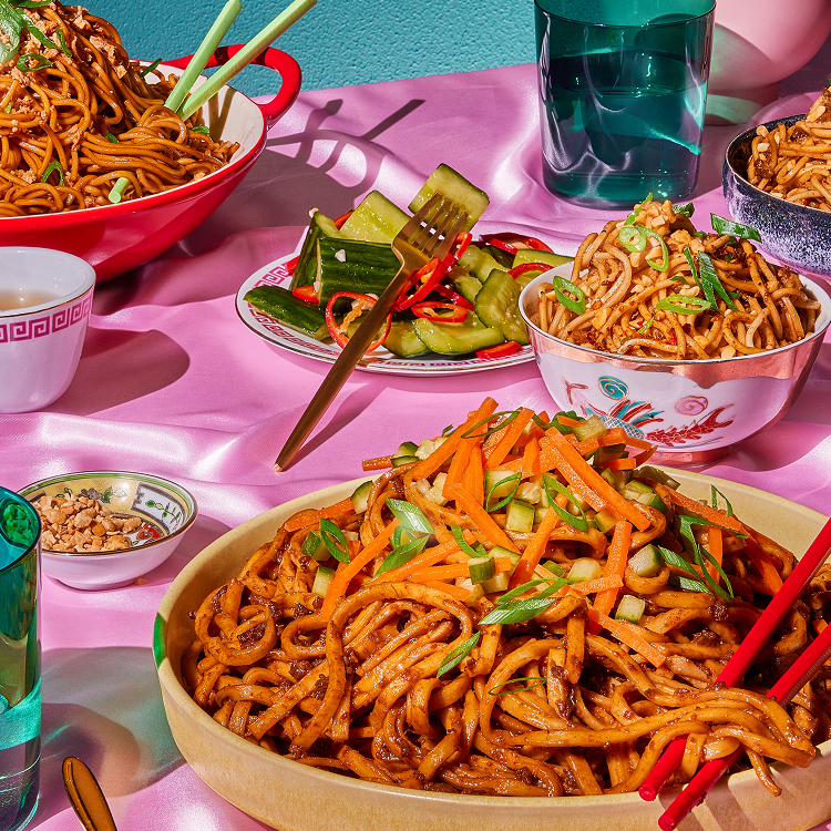 Several bowls of Zha Jiang noodles topped with scallions, served with cucumber salad, crushed peanuts, and red chopsticks on a pink tablecloth