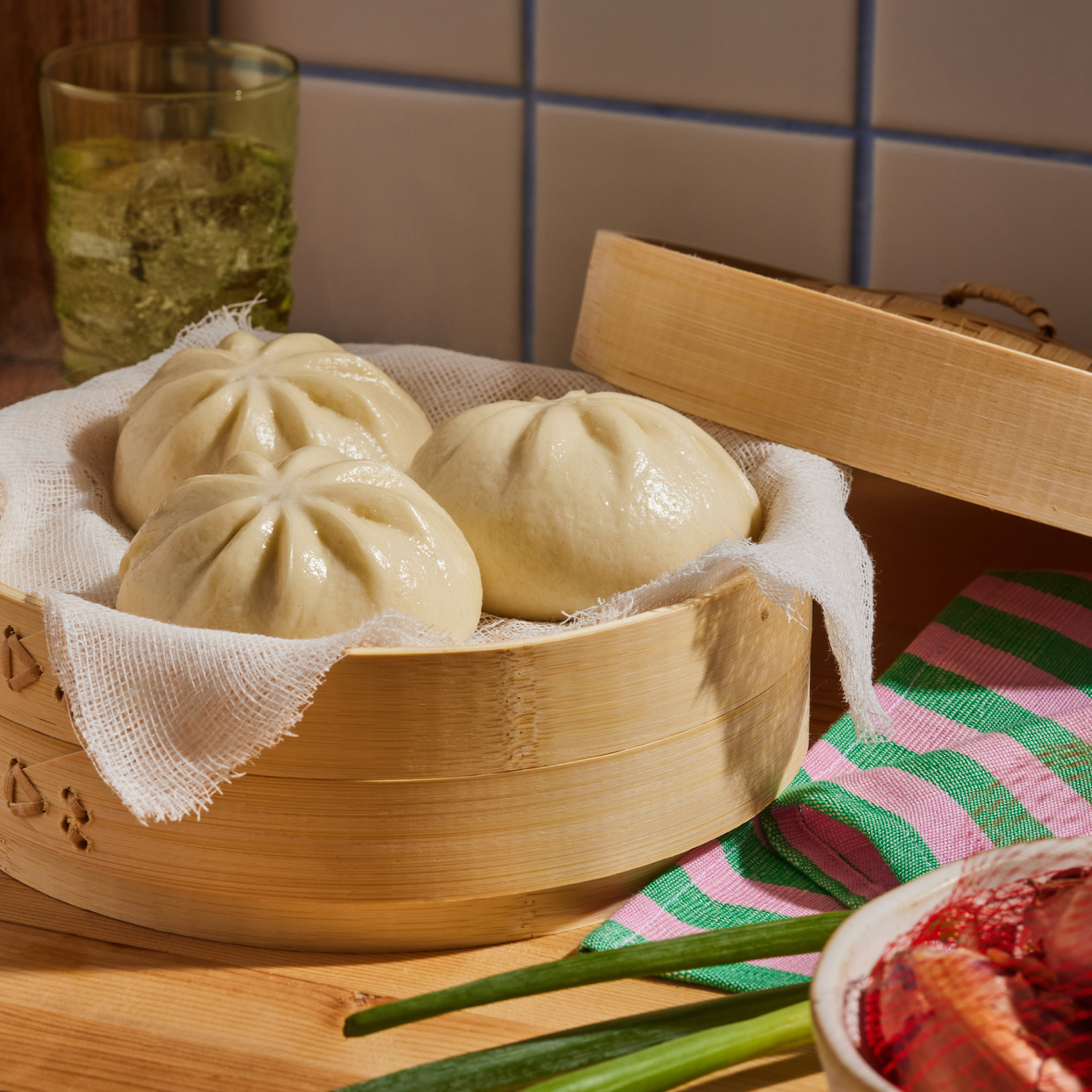 Three steamed Chinese BBQ pork bao buns in a bamboo steamer on a wooden table with scallions and a dipping bowl