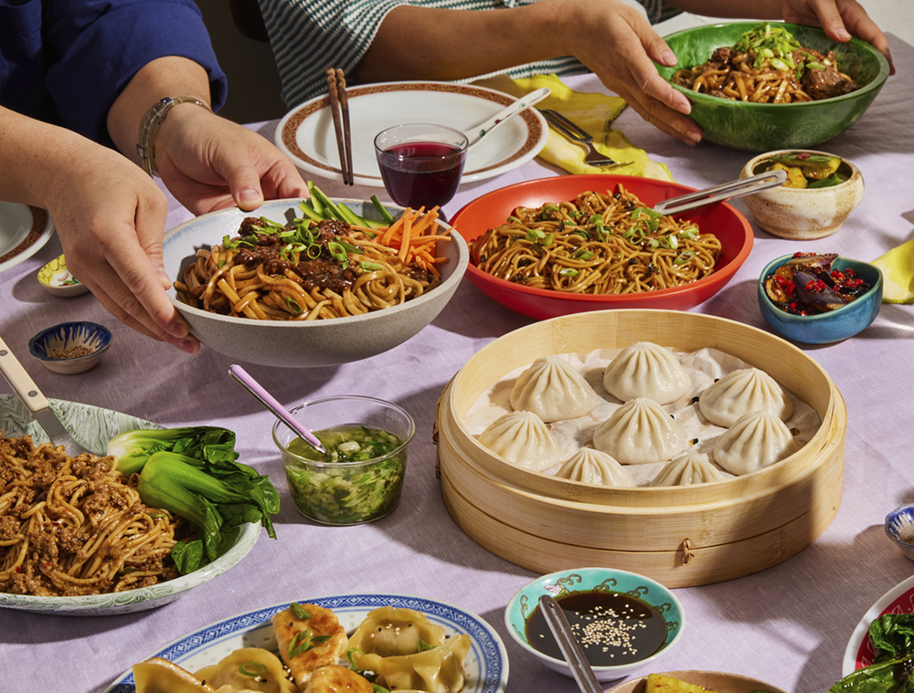 Multiple bowls of noodles, steamed dumplings, and small condiments on a purple tablecloth as people pass a bowl in a family-style meal