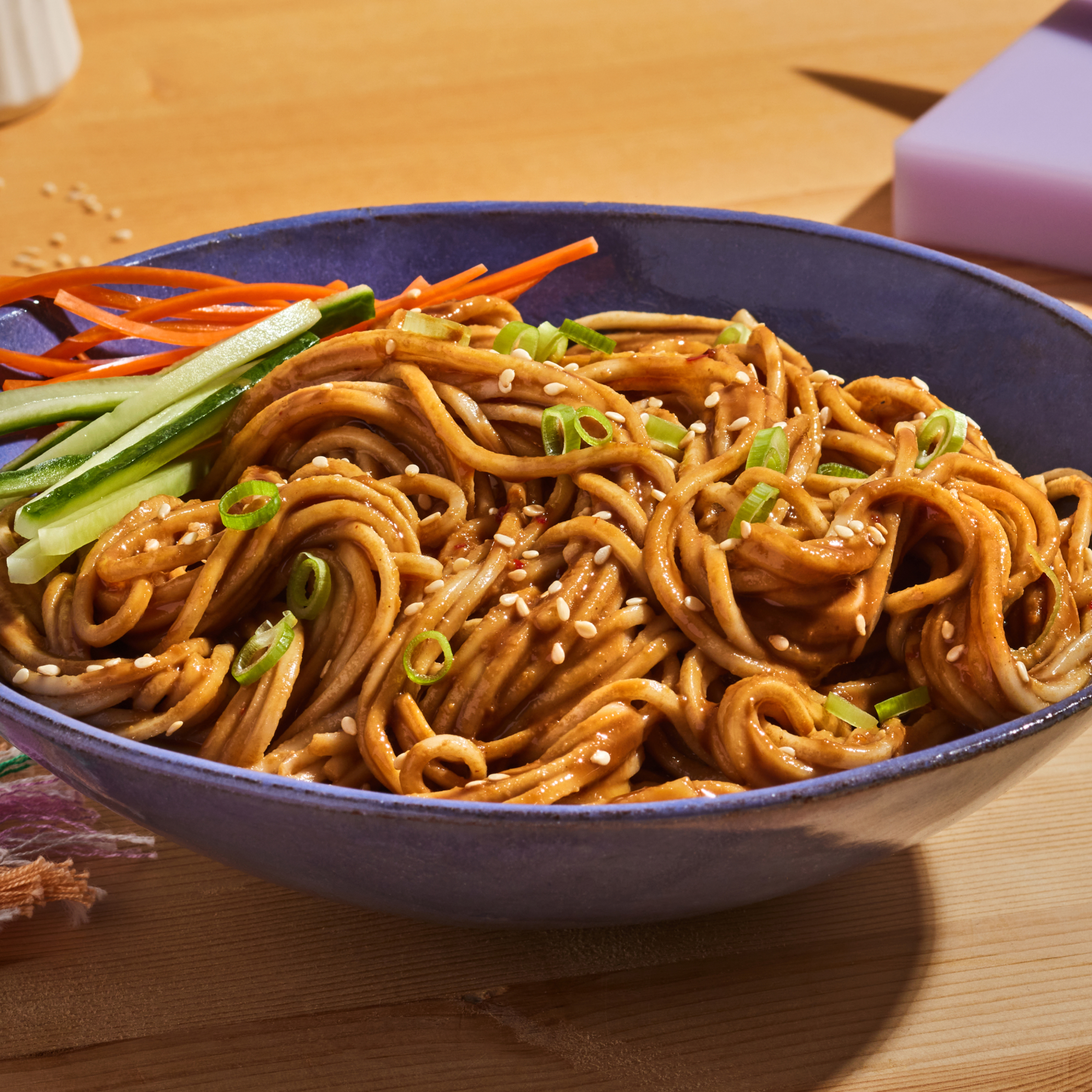Close-up of Creamy Sesame Noodle showing wheat noodles in sesame sauce topped with sesame seeds, sliced scallions and cucumber strips in a blue bowl on a wooden table